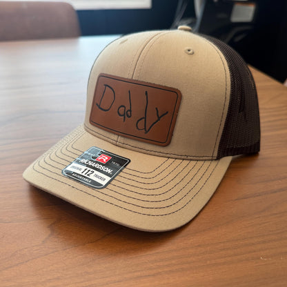 Beige cap with 'Daddy' patch on a wooden surface